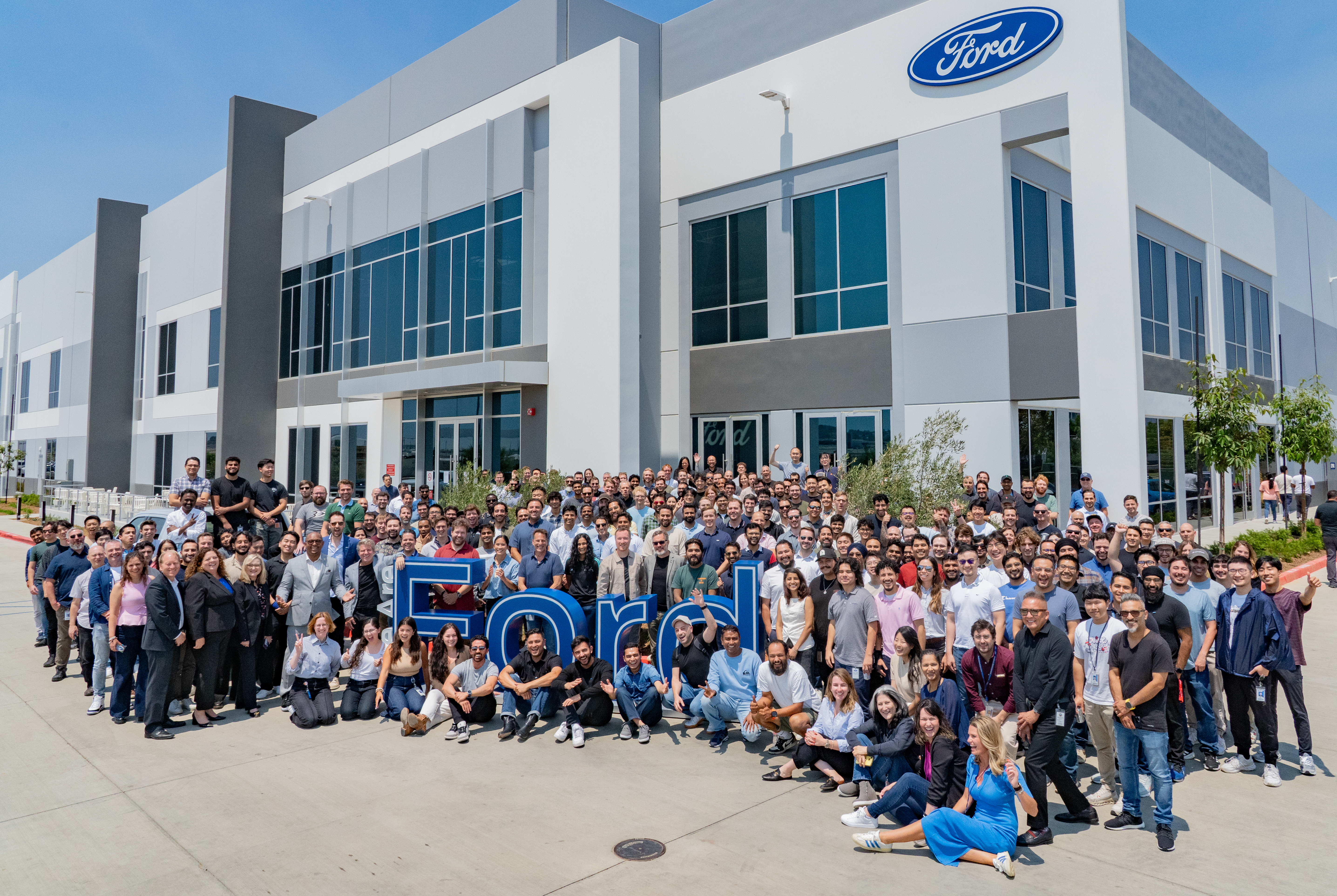 Many employees pose for a photo around large letters spelling out "FORD" in front of a new Ford facility